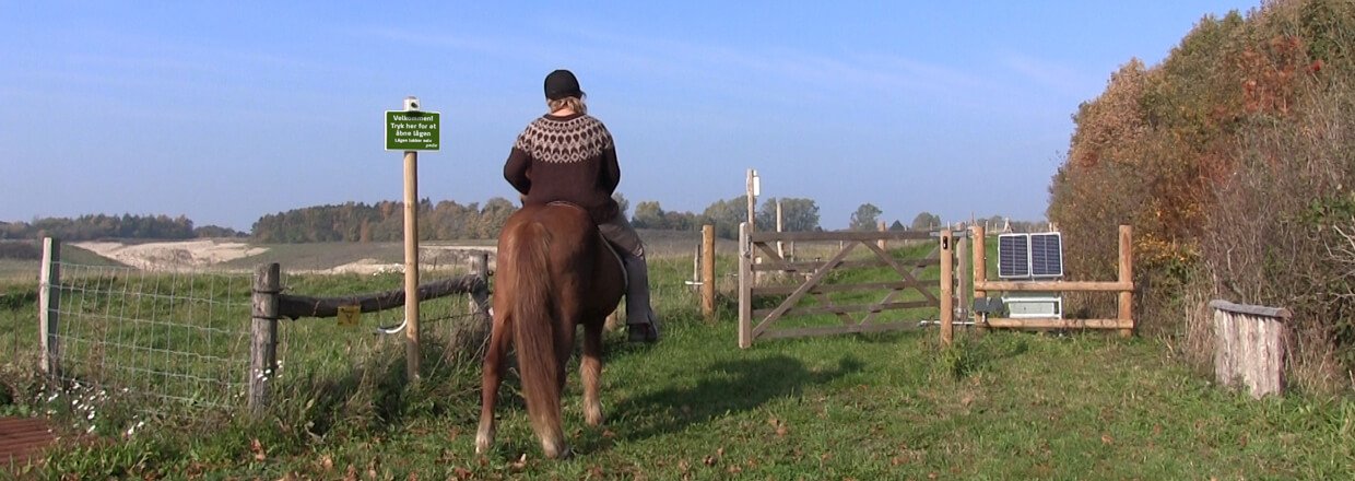 Kvindelig rytter til hest afventer at trælågen foran hendes hest åbnes af en soldrevet lågeautomatik | Poda Hegn